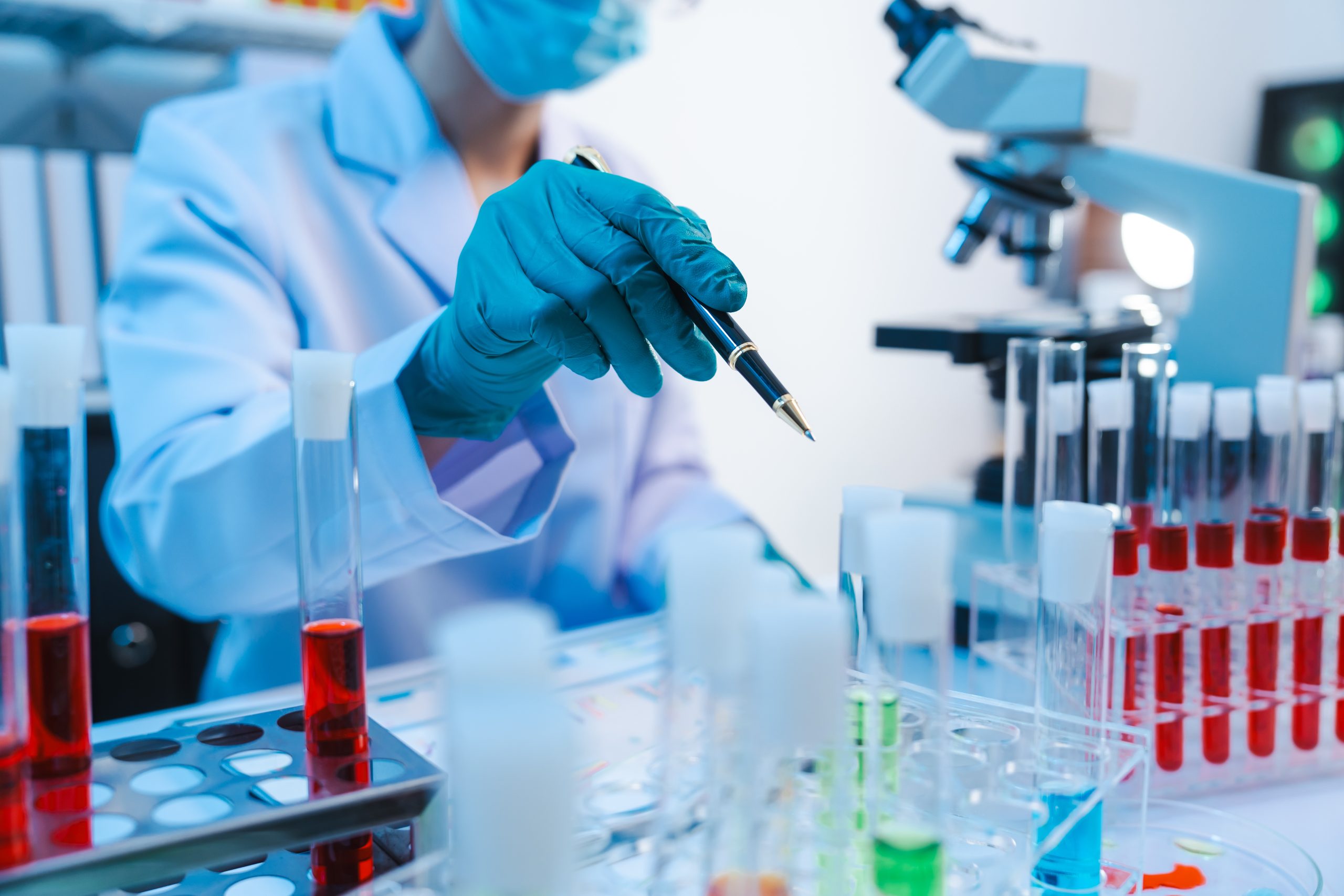 Asian people scientist in lab coat and protective gloves working with test tubes with green and red liquids, with microscope and other test tubes in the background in laboratory.